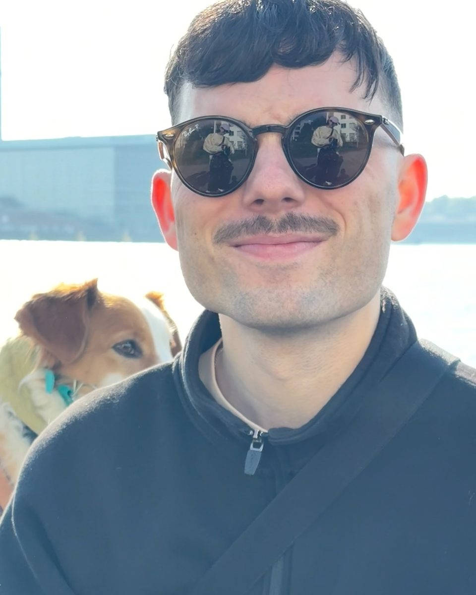 Jakob Rudigier mit Sonnenbrille und Schnurrbart, im Hintergrund ein Hund am Wasser